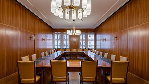 A single wooden chair at a counsel table, papers squared in front of it, the seat beside it empty and pushed in