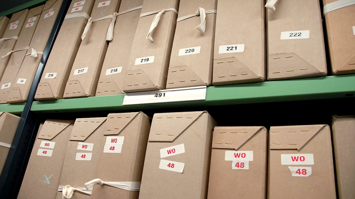 An open cardboard storage box on a concrete floor with loose printed pages stacked unevenly inside it
